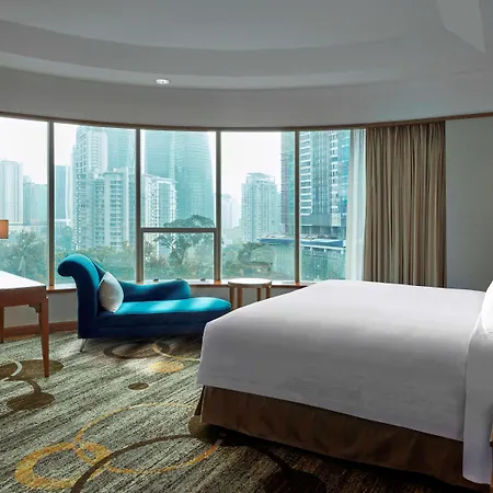 Pullman Kuala Lumpur City Centre Hotel & Residences Exterior photo The photo shows a hotel room featuring a large window with a view of city skyscrapers. The room includes a neatly made bed with white linens, a blue lounge chair, a wooden desk with a chair, and a lamp on a side table. The flooring has a patterned ca